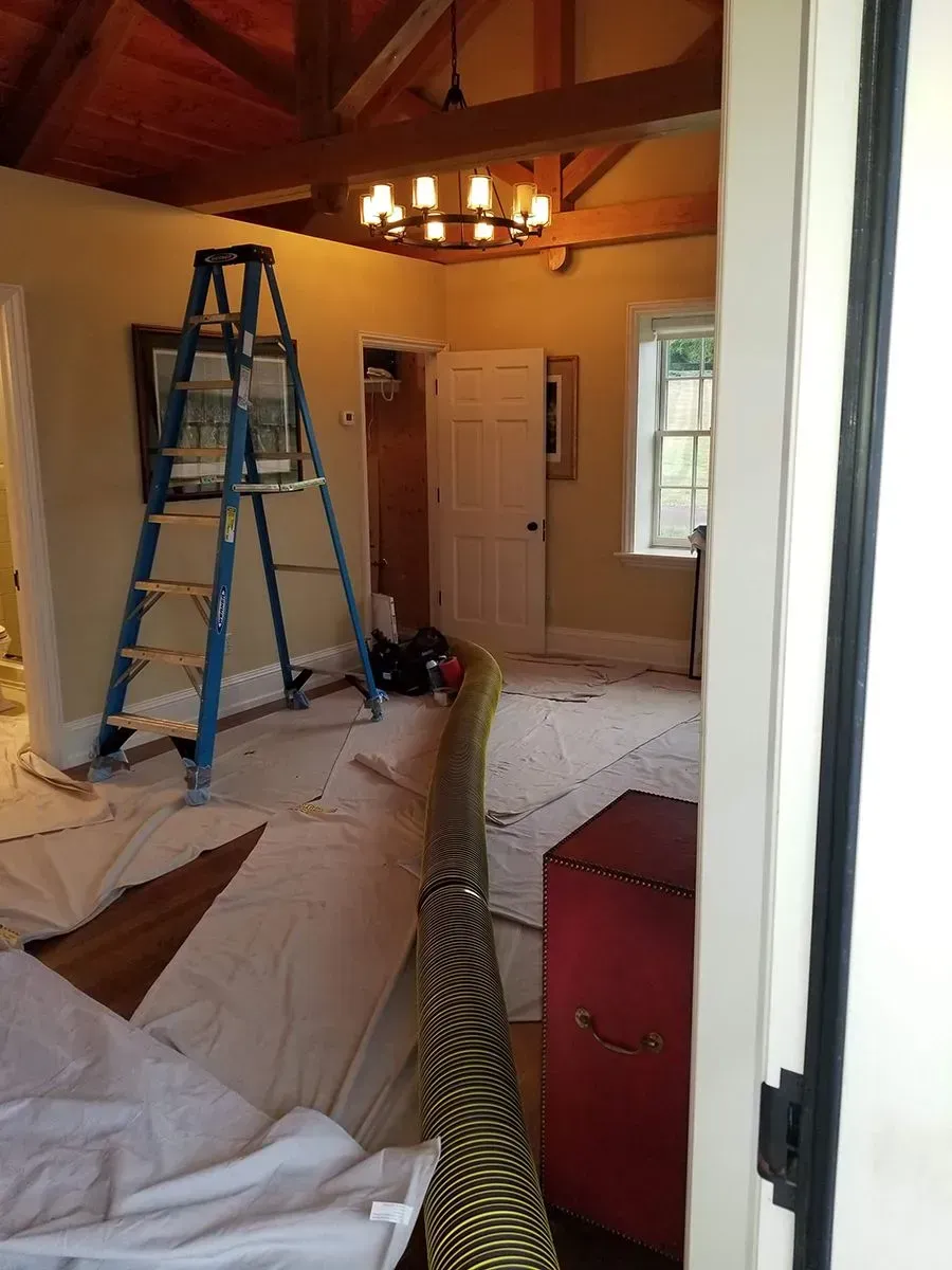 Room being painted, with a ladder, drop cloths, and red trunk. Beige walls and wooden ceiling beams.