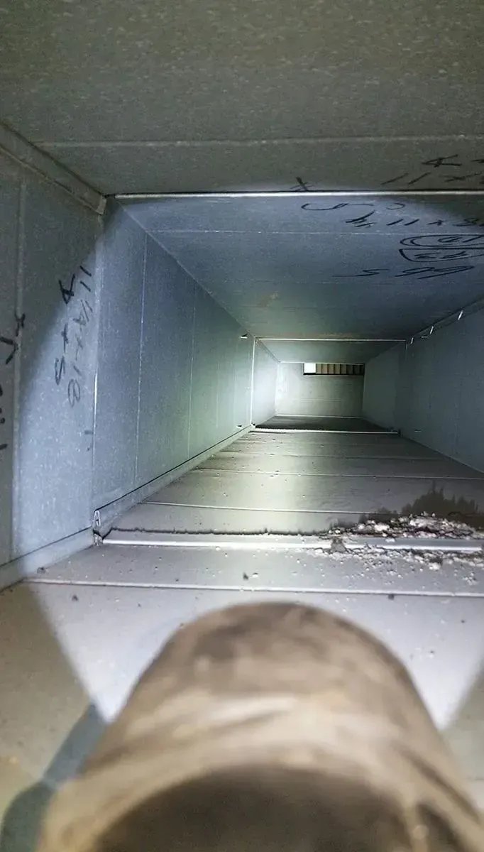 Interior of a rectangular duct, illuminated by a flashlight. Gray metal walls. Debris on floor.