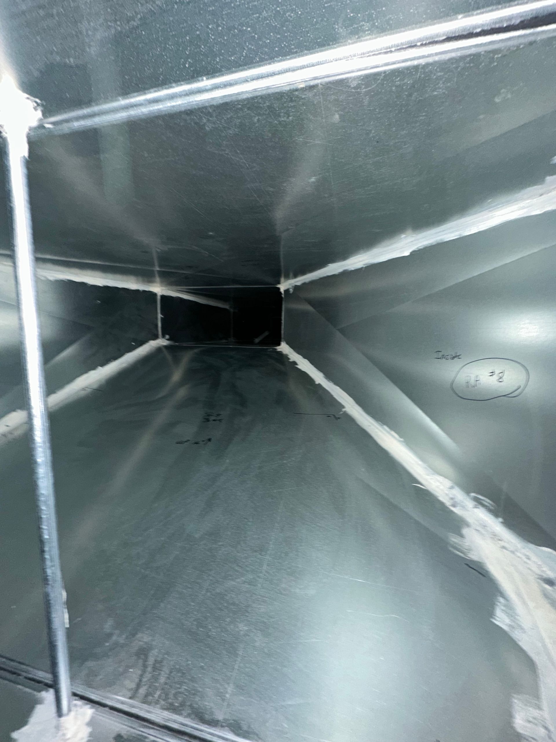 Inside view of a rectangular metal duct. White sealant lines seams, leading to a dark opening.