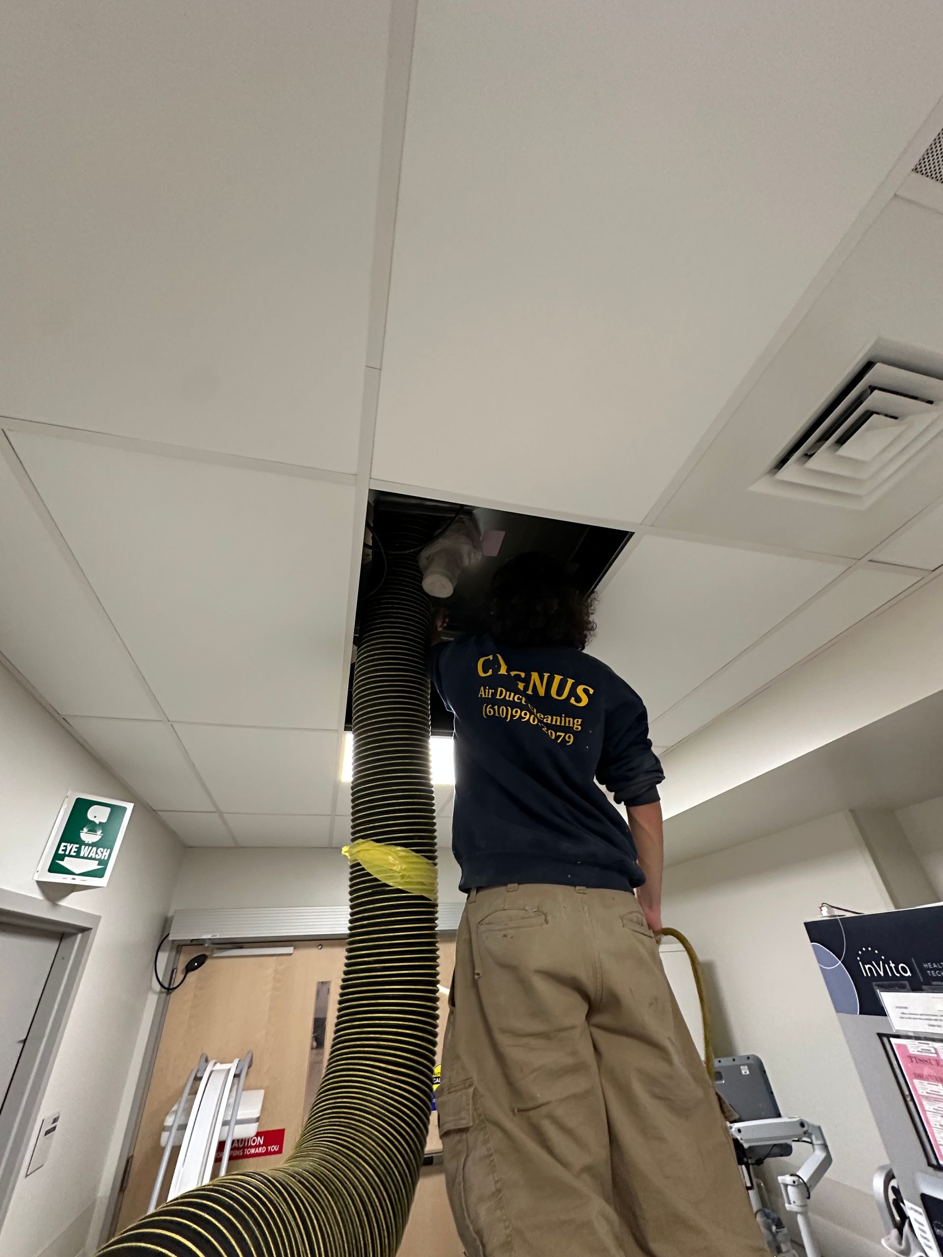 Person on ladder working on ceiling; large black hose in open ceiling panel. Interior setting.