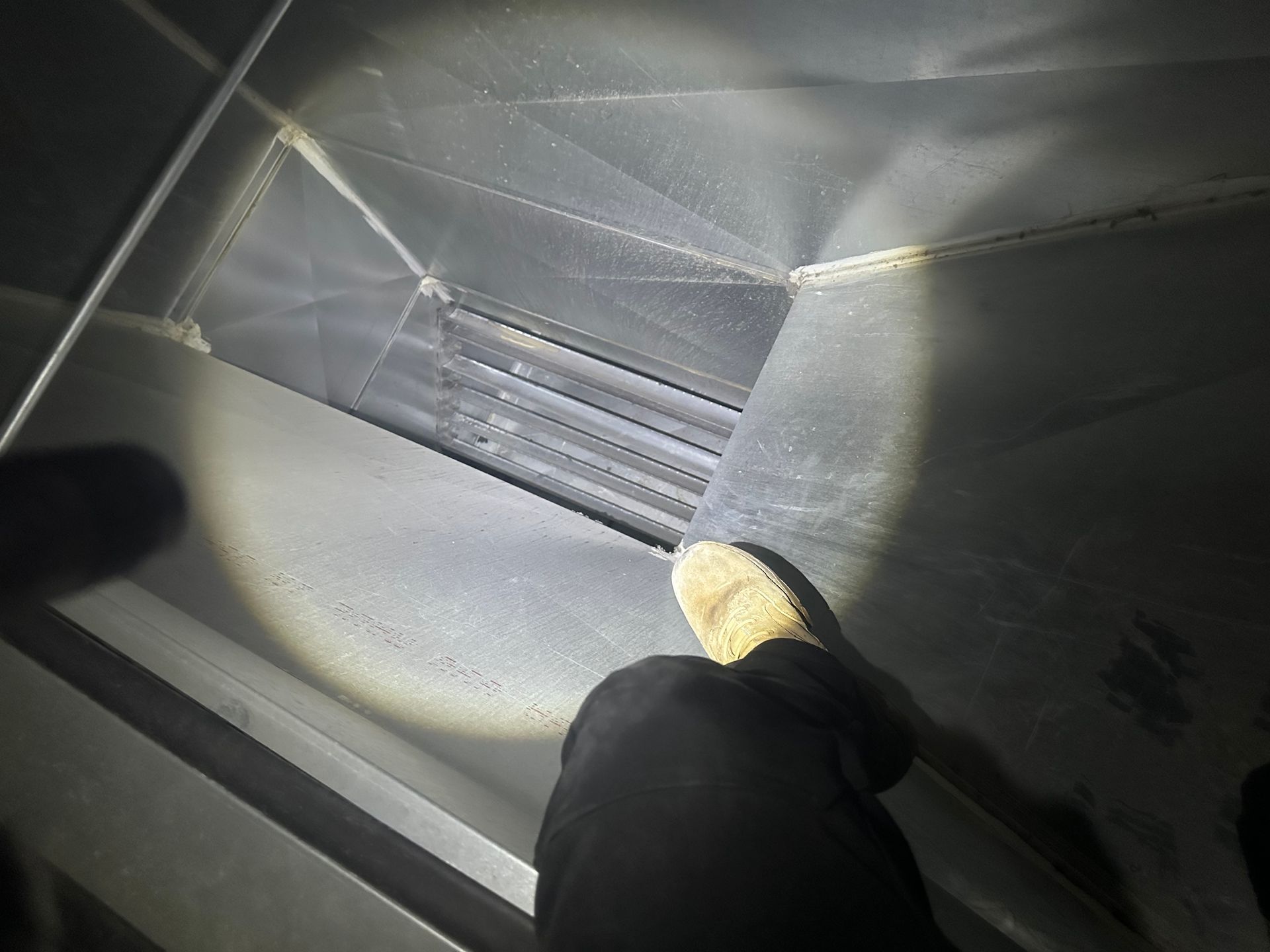 Person's foot in ductwork, illuminated by a flashlight. Metal duct interior with vents and seams visible.