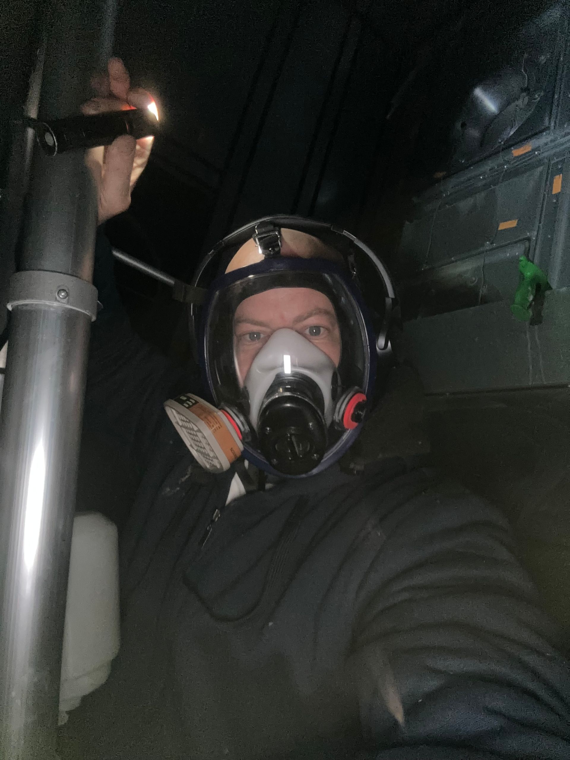 Person in respirator mask, holding a flashlight in a dark space, near a metal pipe.