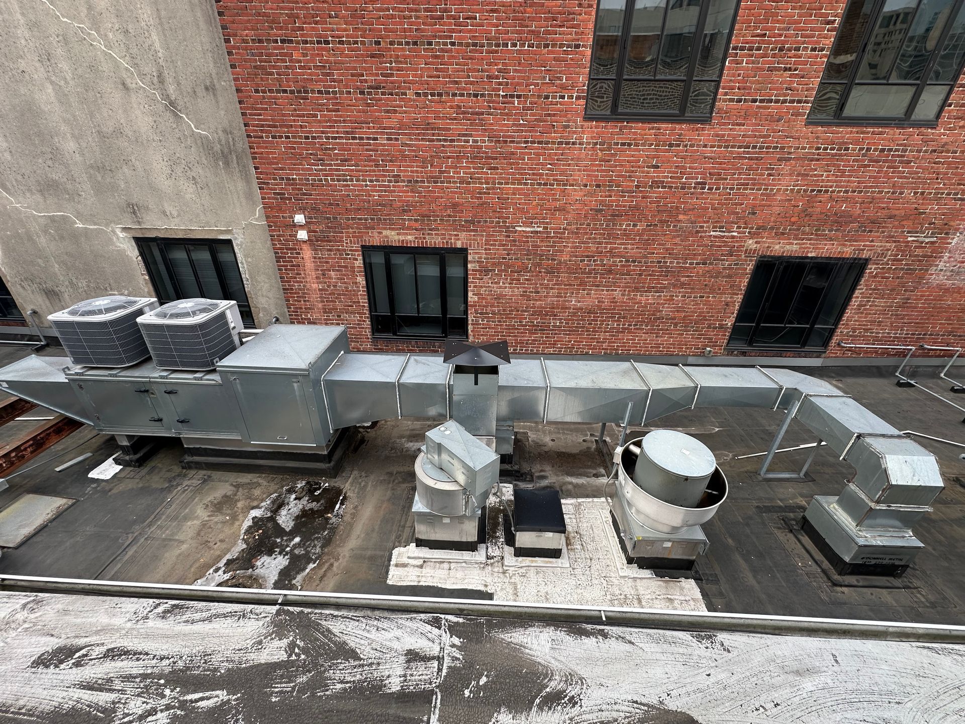 Rooftop with HVAC units and ductwork against brick building with windows. Snow covers parts of the surface.