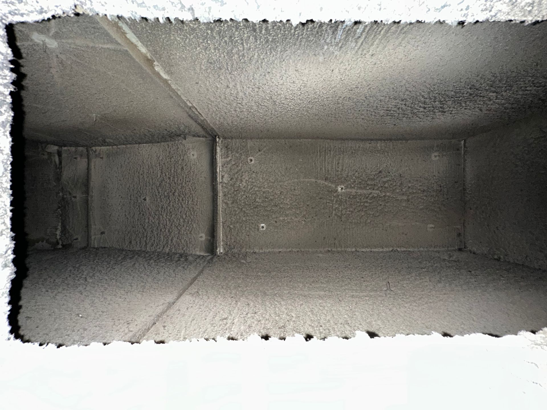 Interior view of a rectangular, dusty air duct, coated in gray grime.