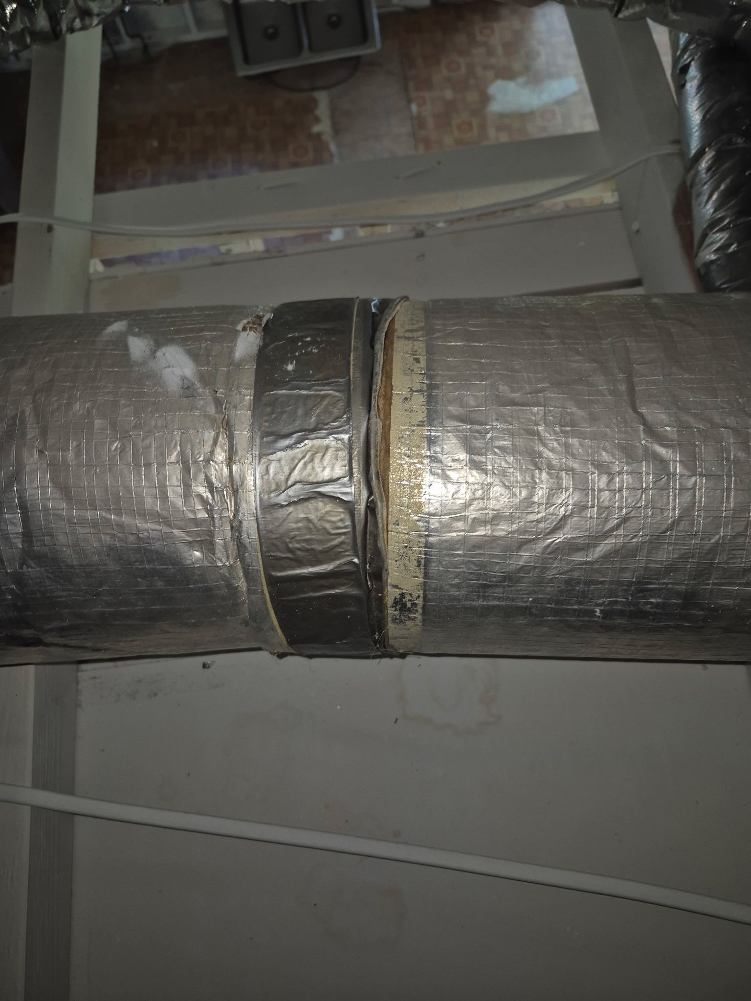 Silver insulated ducting, joined with black tape, above a white wall.
