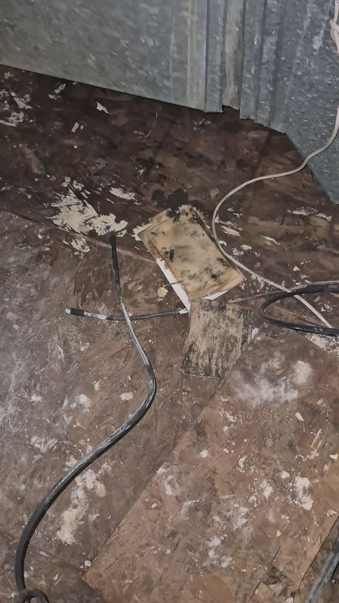 Damaged floor with exposed wood, wires, and debris in a room with a metal wall.