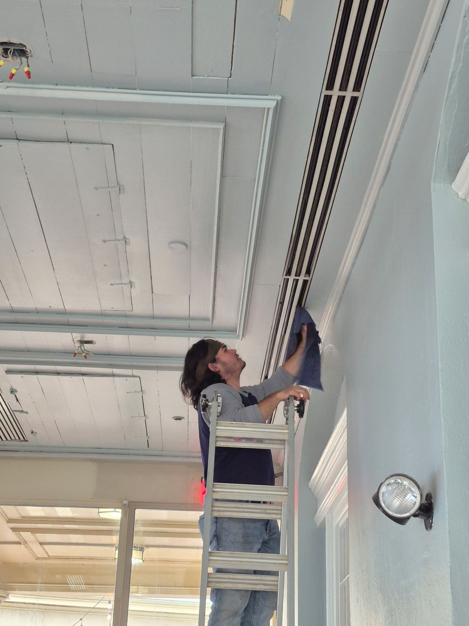Person on a ladder, installing something on a ceiling with striped design. Light blue wall and molding visible.