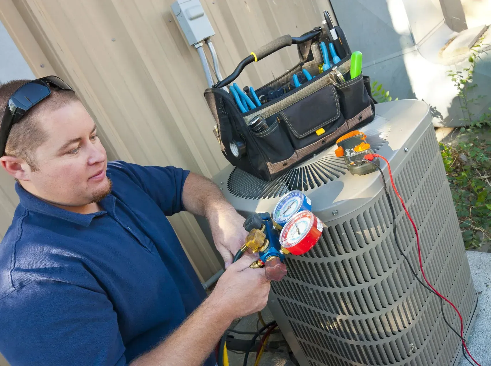 HVAC technician working on an air conditioning unit outside, using tools and gauges.