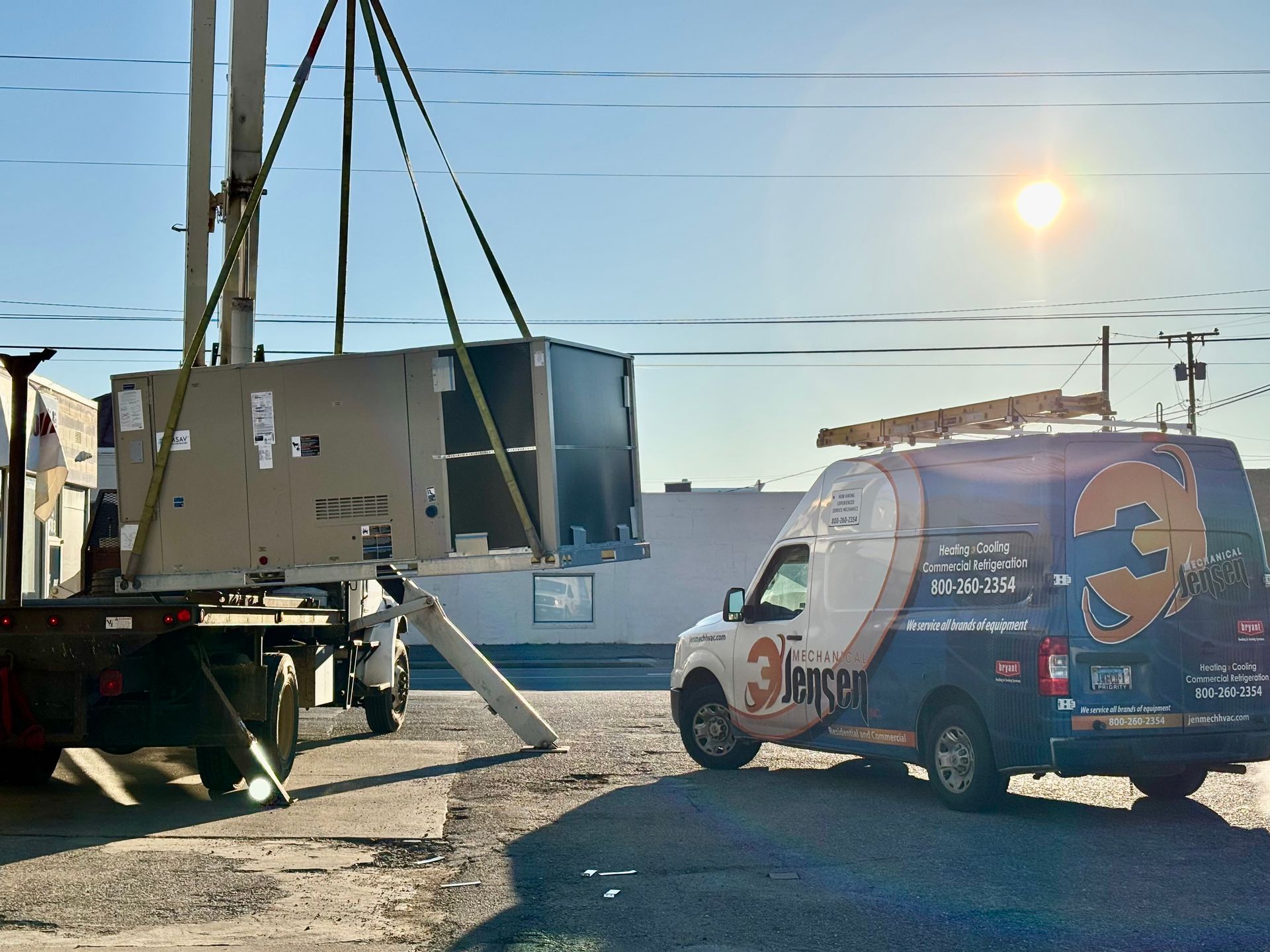 A large HVAC unit being lifted by a crane off a truck, near a service van on a sunny day.