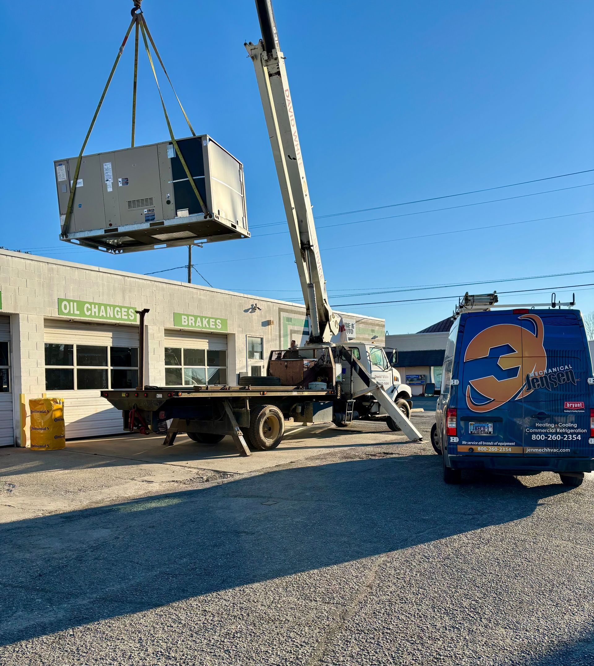 A crane lifts a large HVAC unit above a building. A service van is parked nearby.