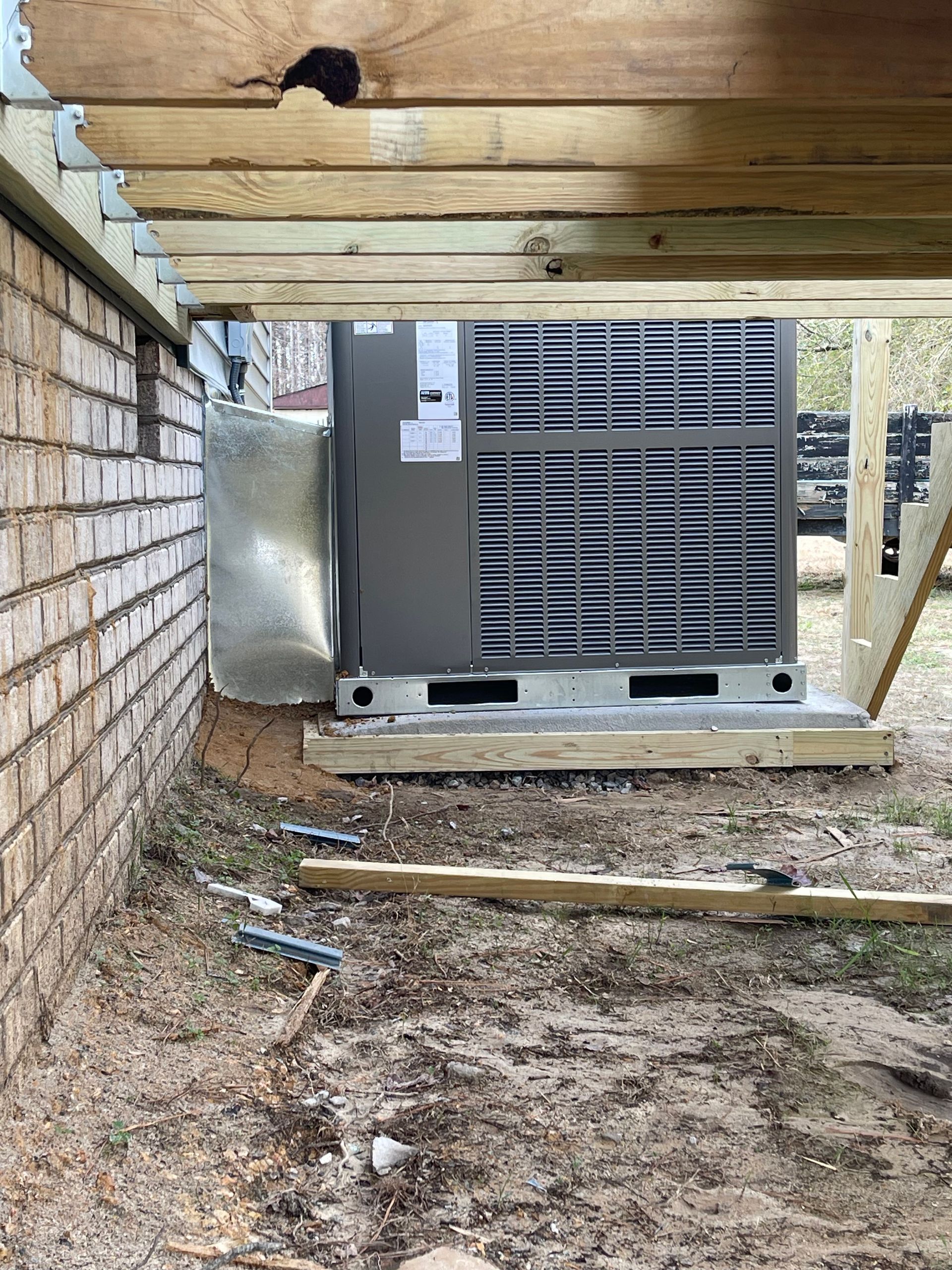 An air conditioning unit on a wooden base under a wooden deck, next to a brick wall.