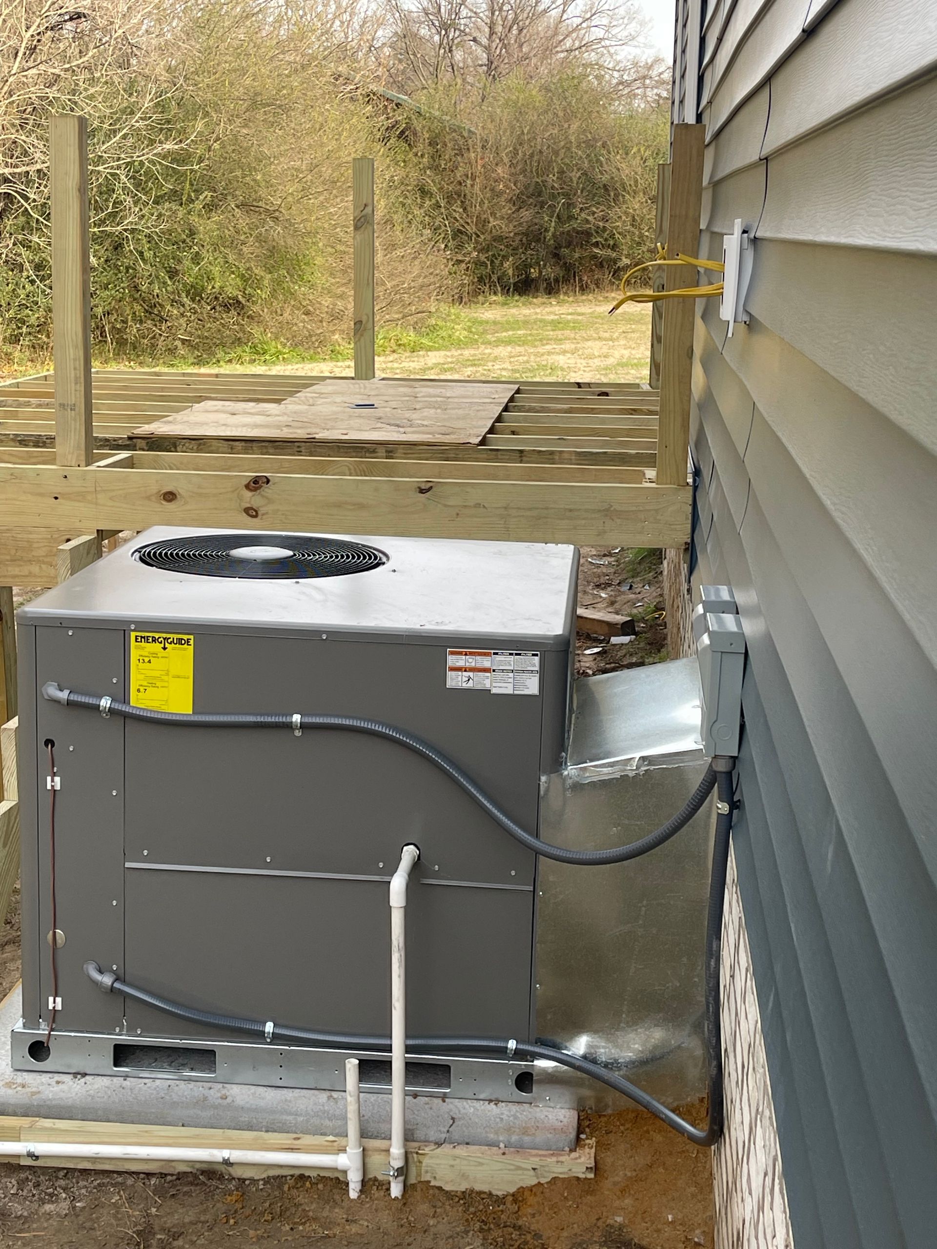 AC unit on concrete base next to a building. Wiring connects unit to a wall-mounted box. Wooden deck in the background.
