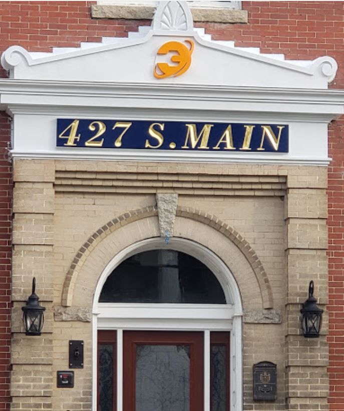 Building entrance at 427 S. Main with sign, orange logo, brick and beige facade, arched doorway, and sconces.