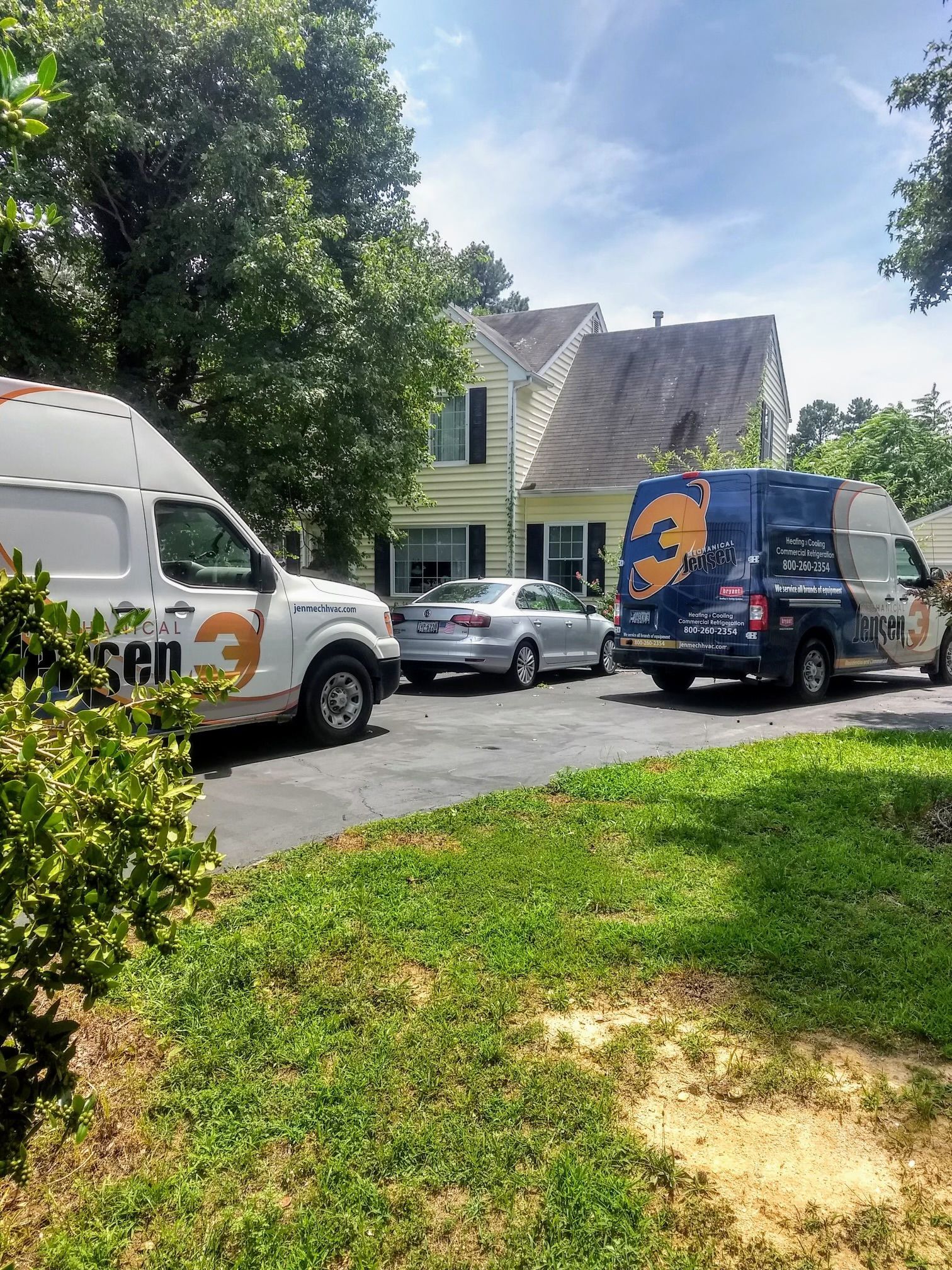 Two work vans parked in a driveway, in front of a yellow house with a silver car parked between them.