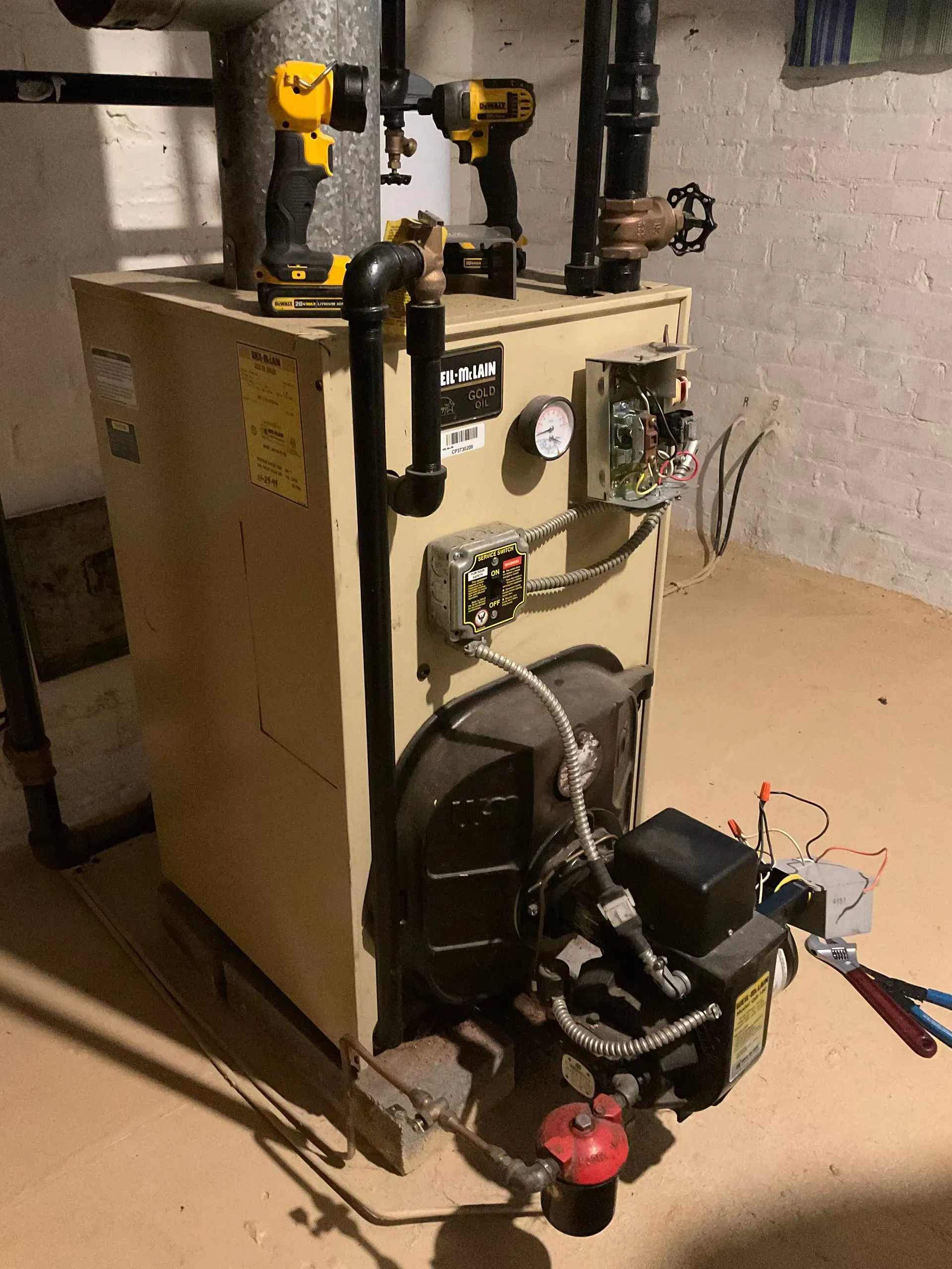 Tan furnace in a basement setting with black pipes and exposed electrical components.