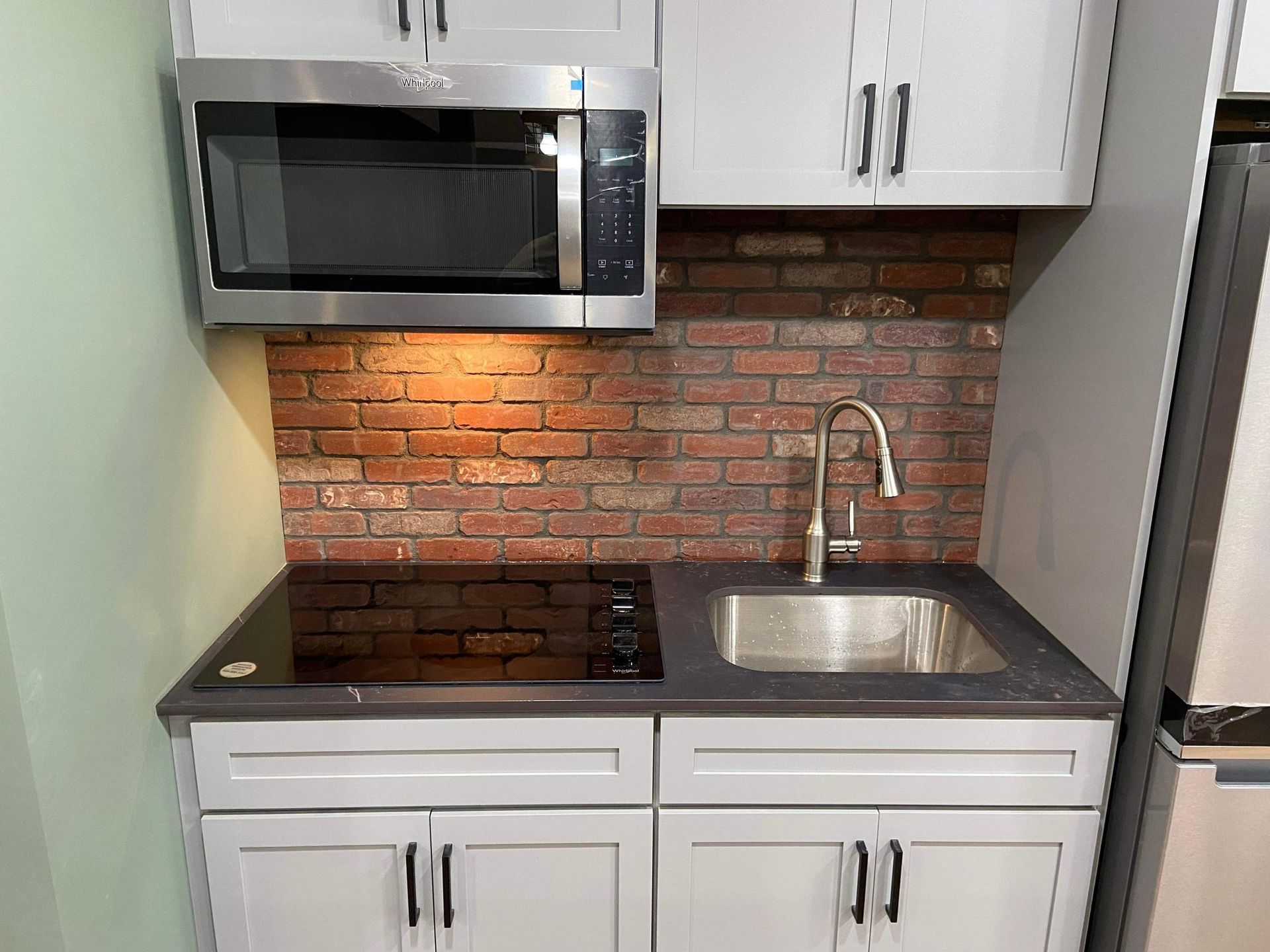 A kitchen with white cabinets , a sink , a microwave , and a brick wall.