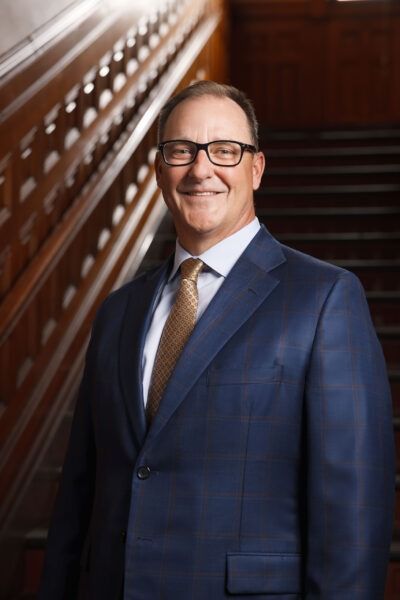 Smiling person in a navy suit and glasses standing on a staircase in a wood-paneled interior