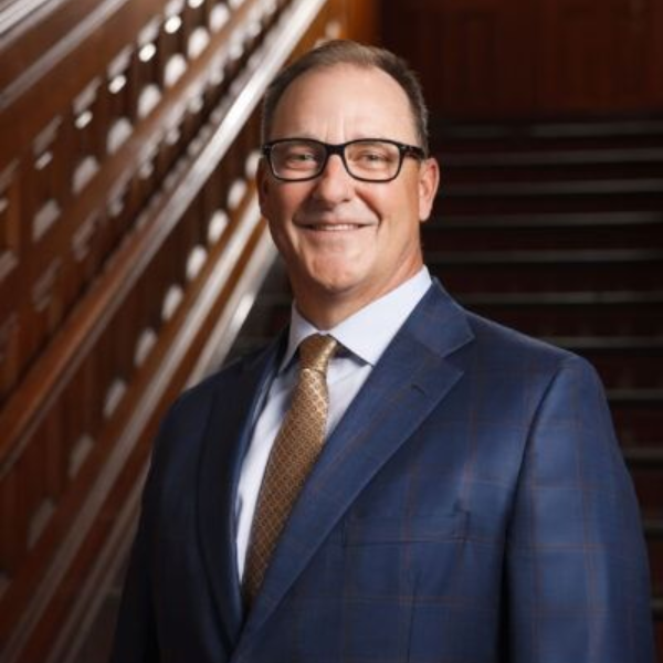 Smiling person in a navy suit and glasses standing on a staircase in a wood-paneled interior