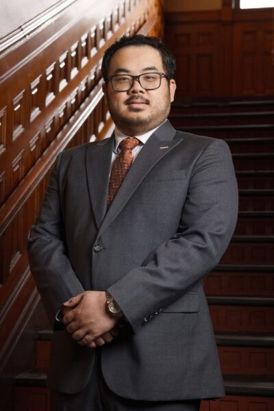 Man in a gray suit standing on a staircase with wooden railings, hands clasped in front of him.