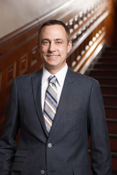 Man in a dark suit and striped tie standing on a staircase, smiling at the camera.