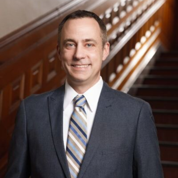 Man in a dark suit and striped tie standing on a staircase, smiling at the camera.