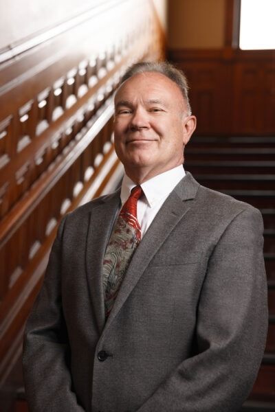 Man in gray suit standing on a wooden staircase, smiling in a formal interior.