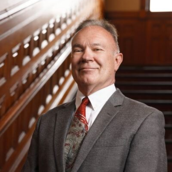 Man in gray suit standing on a wooden staircase, smiling in a formal interior.