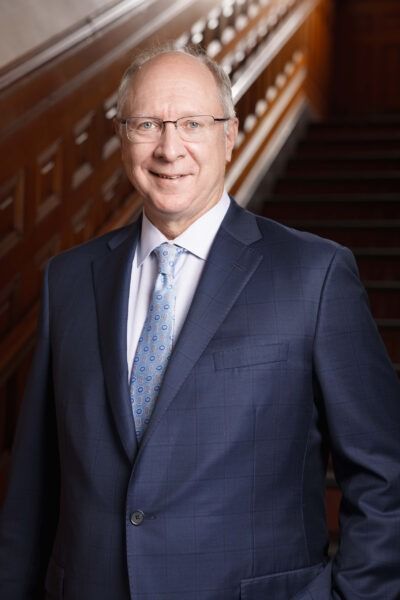 Man in a navy suit standing by a staircase, smiling at the camera