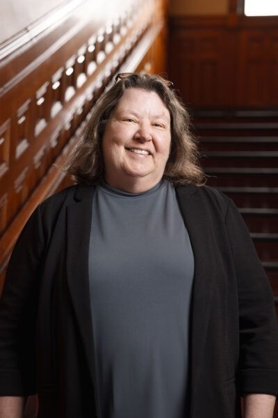 Smiling person in a gray dress and black cardigan standing on a staircase.