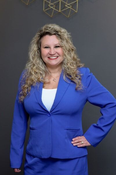 Woman in blue blazer smiling with hand on hip against a dark gray background