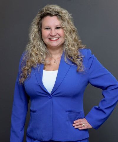 Woman in blue blazer smiling with hand on hip against a dark gray background