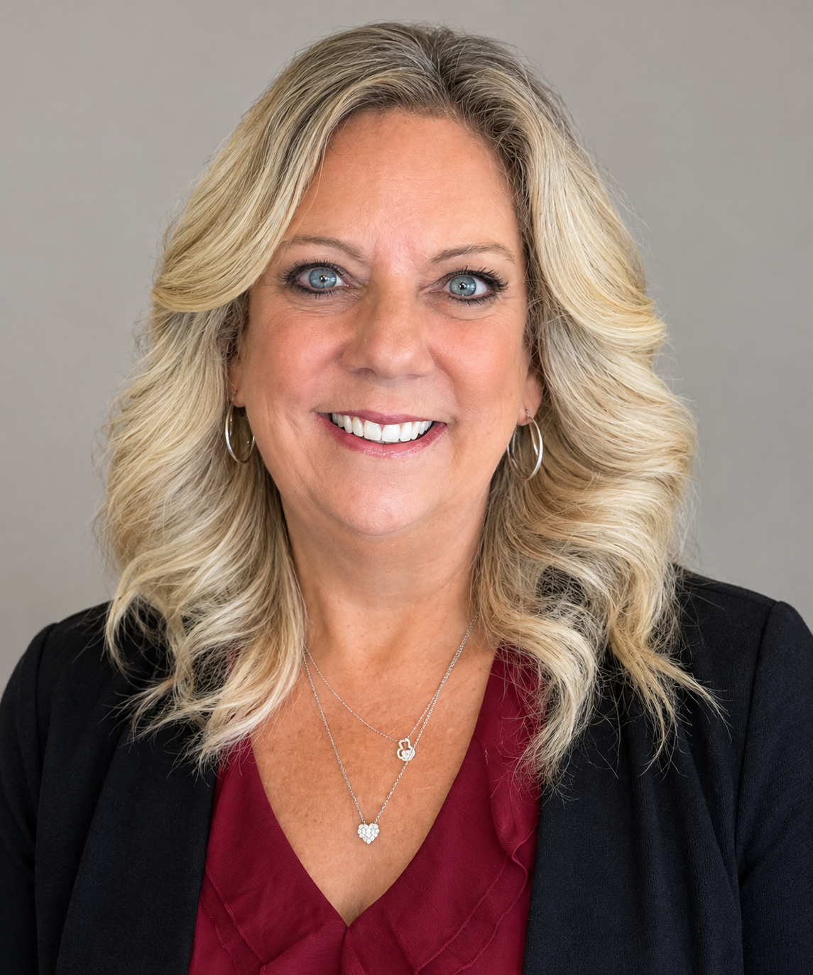 Portrait of a smiling woman with blonde hair wearing a black blazer and burgundy top against a gray background
