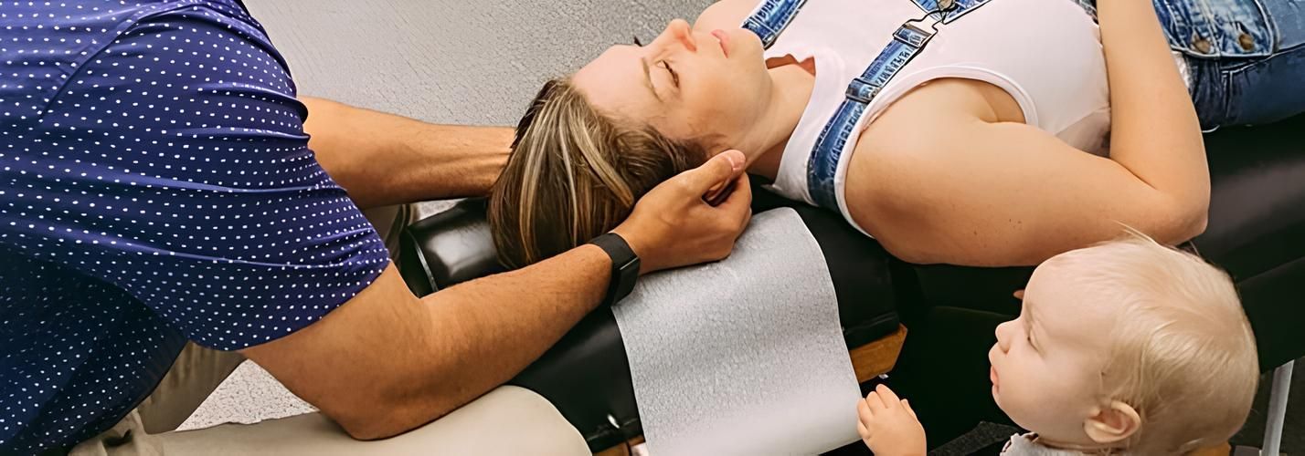 A woman receiving chiropractic care while her baby watches. The woman wears denim overalls.