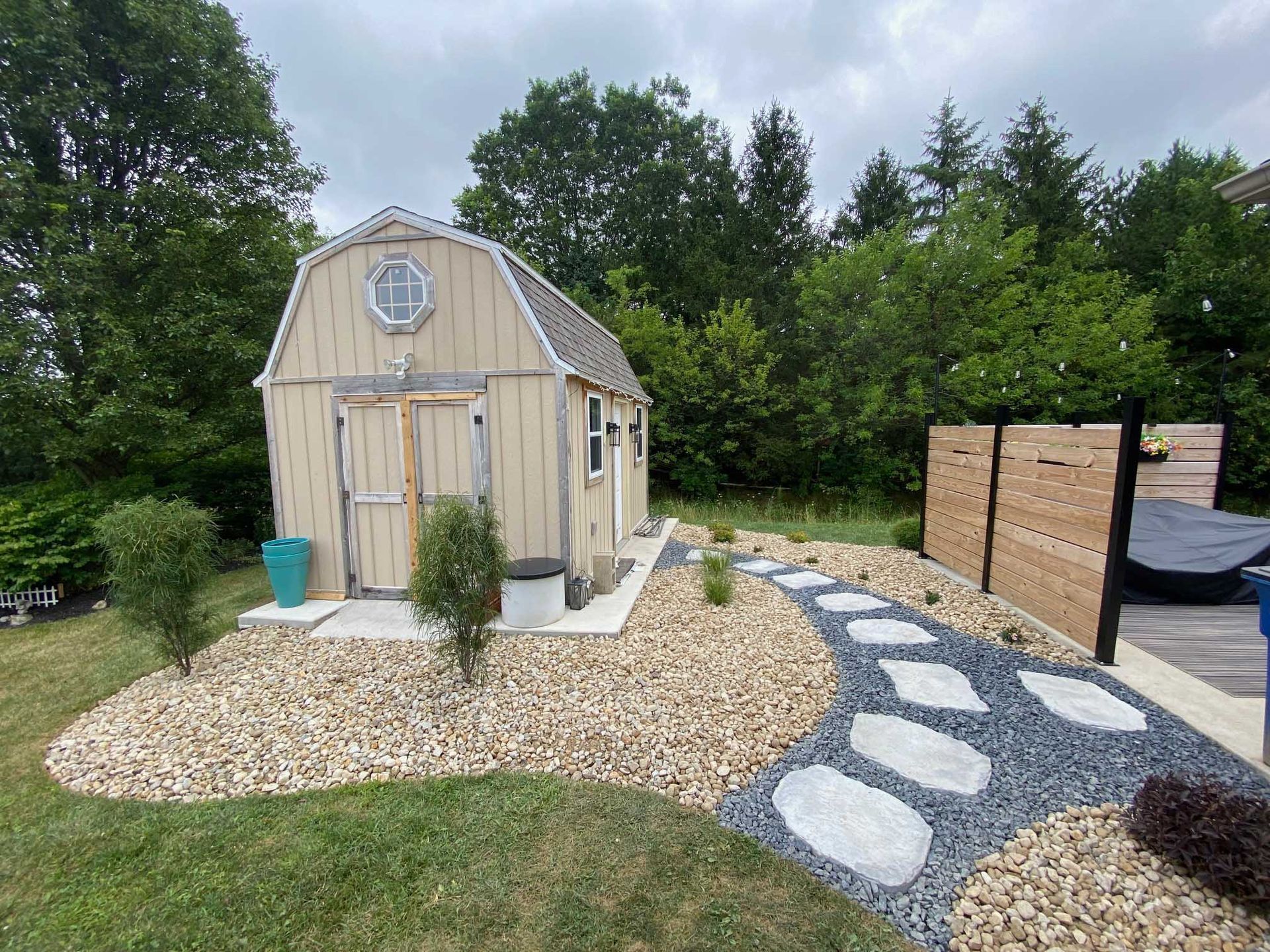 Tan shed in a yard with a rock pathway and landscaping. Trees and a fence are in the background.