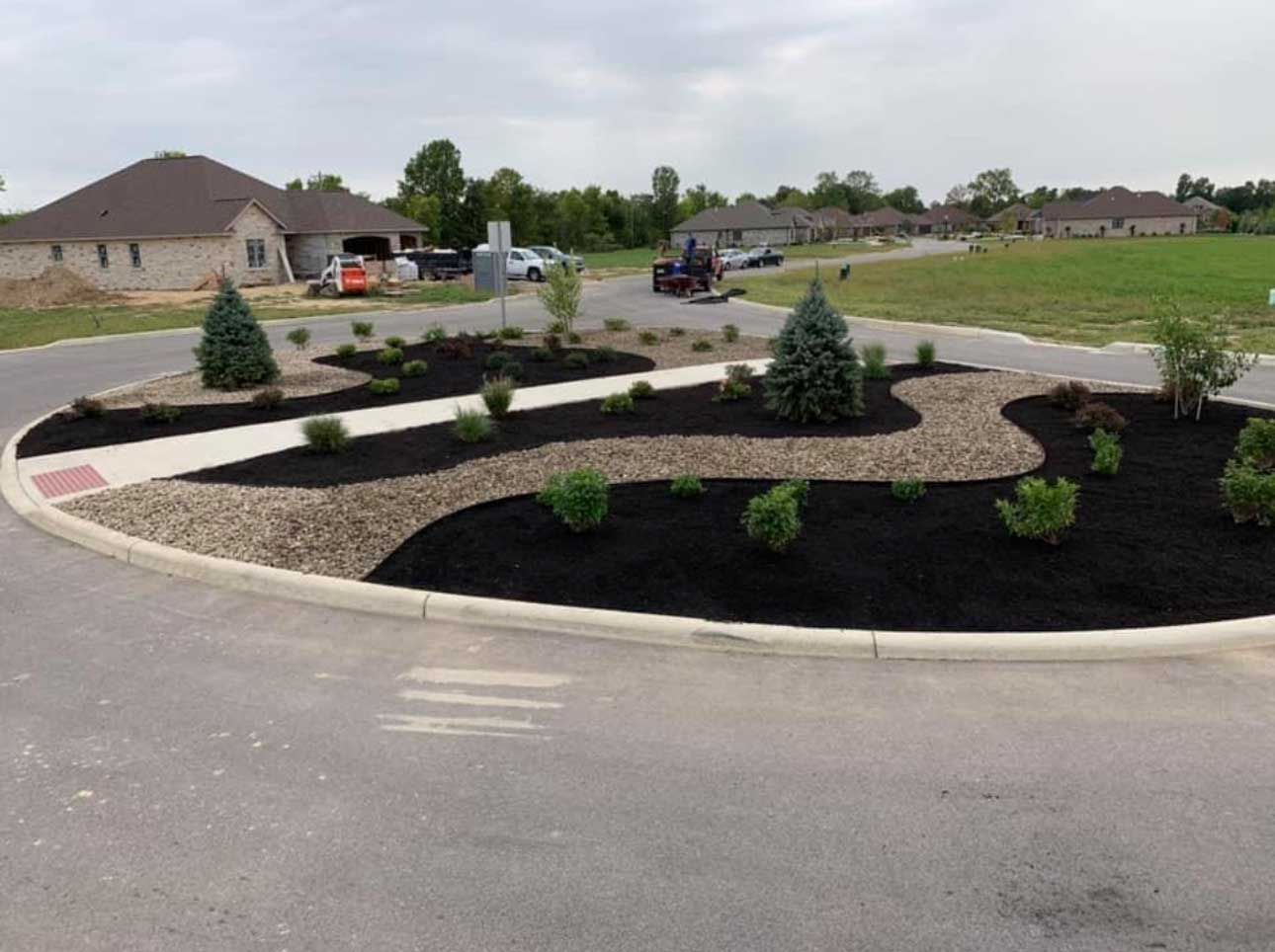 Landscaped roundabout with plants, dark mulch, stone, and a walkway in a residential area.