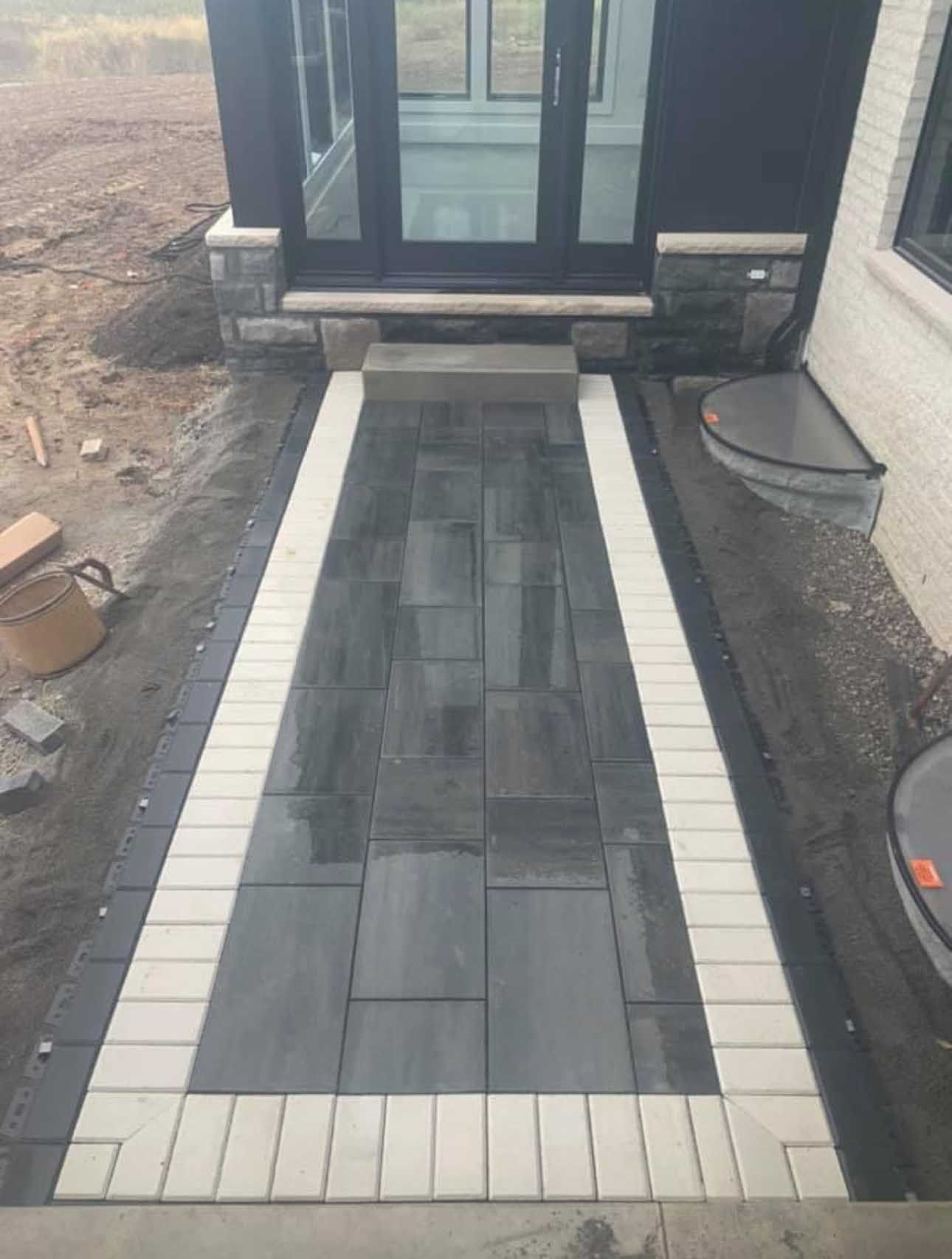 Exterior pathway of dark pavers bordered by light pavers, leading to a doorway.