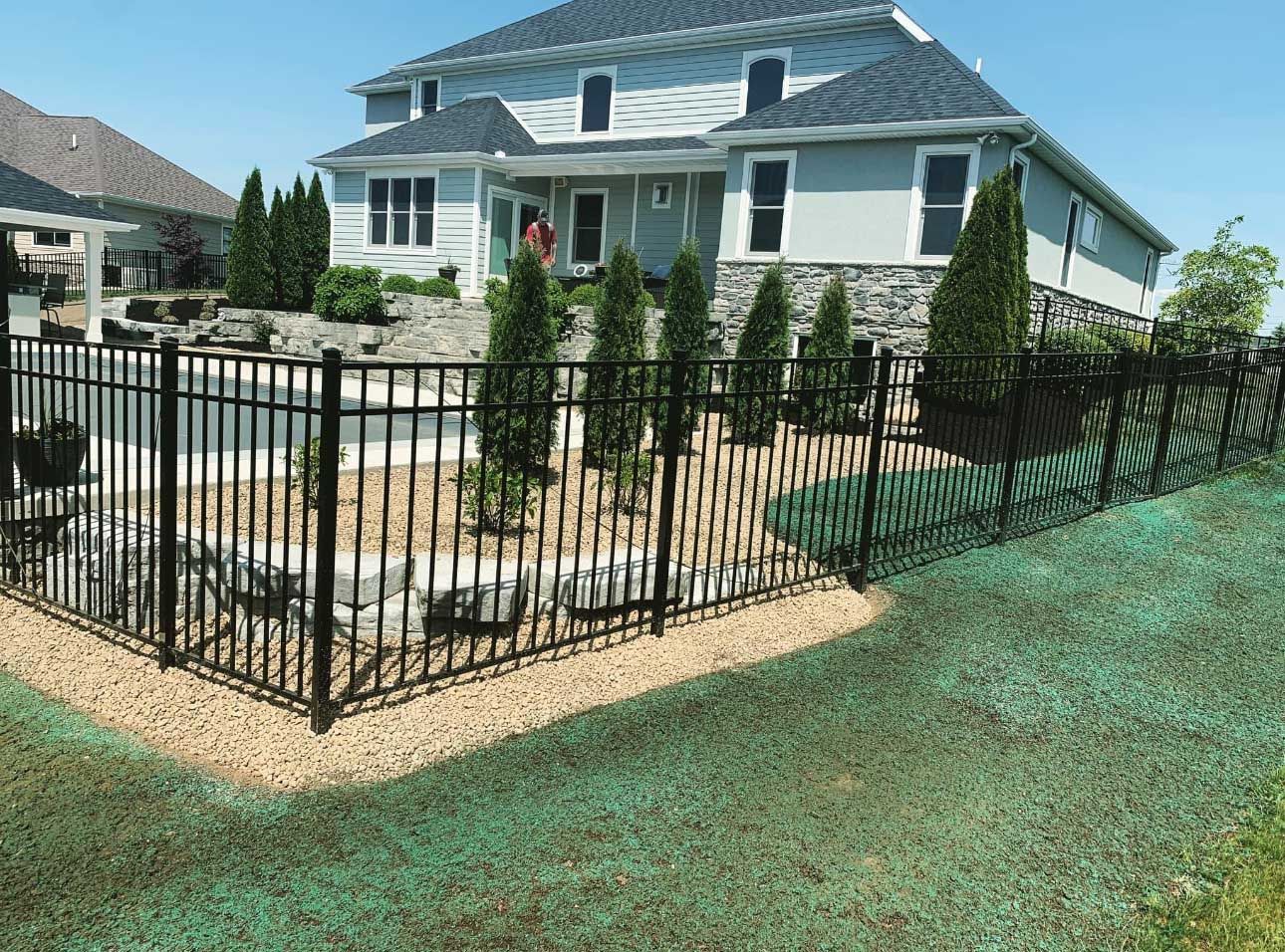 Black fence surrounds a pool in front of a light blue house on a sunny day.