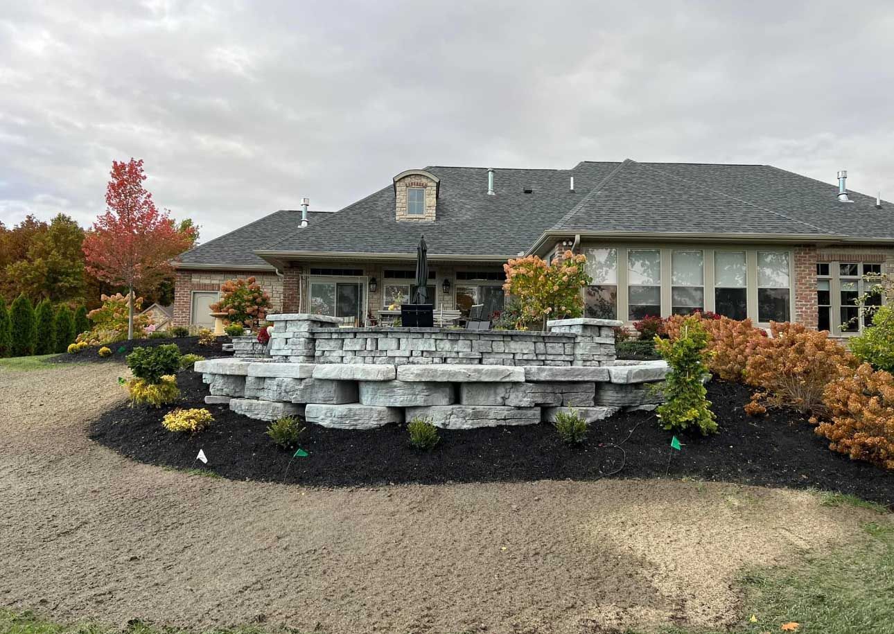 Stone patio with landscaping in front of a brick house with dark gray roof, under a cloudy sky.