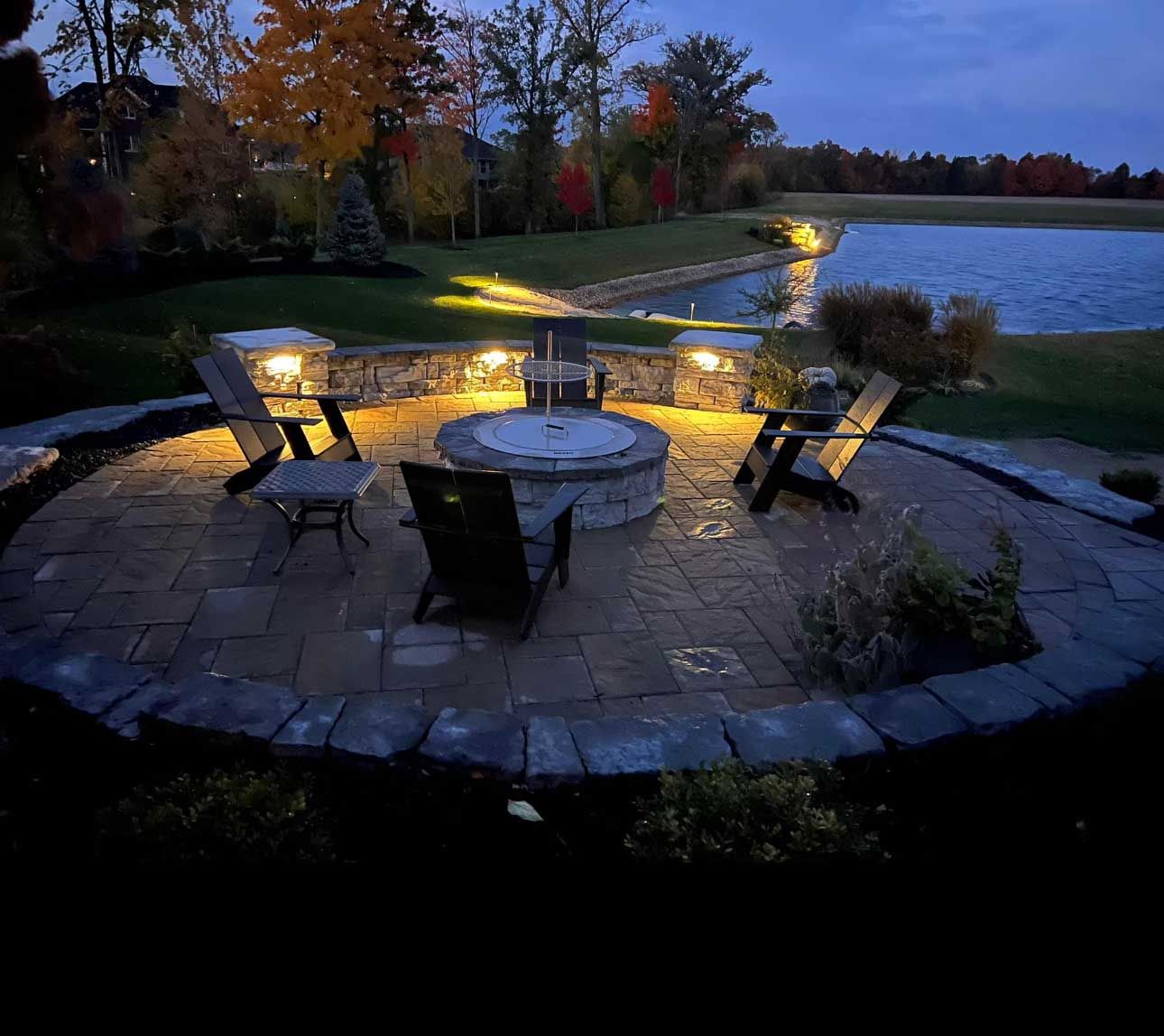 Nighttime outdoor patio with fire pit, seating, and lake in background. Warm lights illuminate the area.