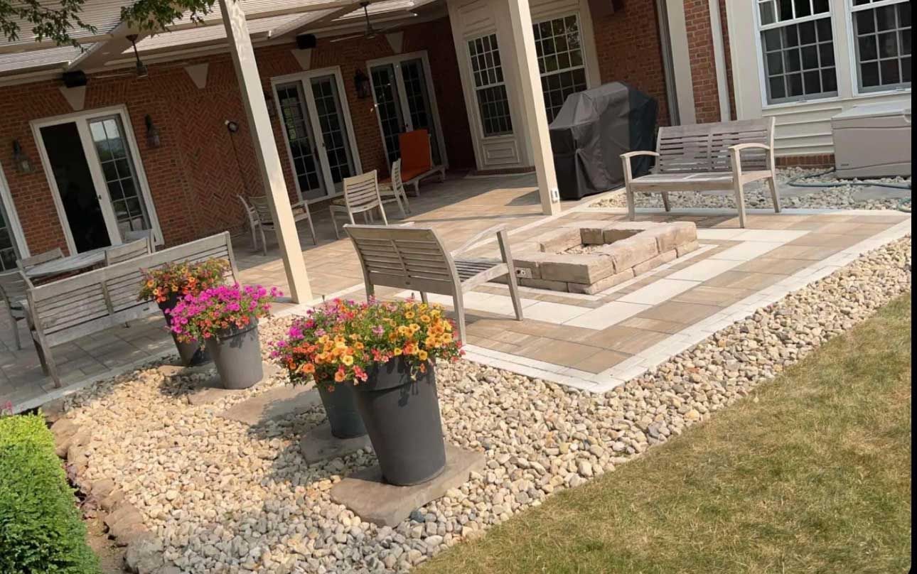 Patio with a fire pit, seating, and flower pots. Beige brick patio with stone border and grass.