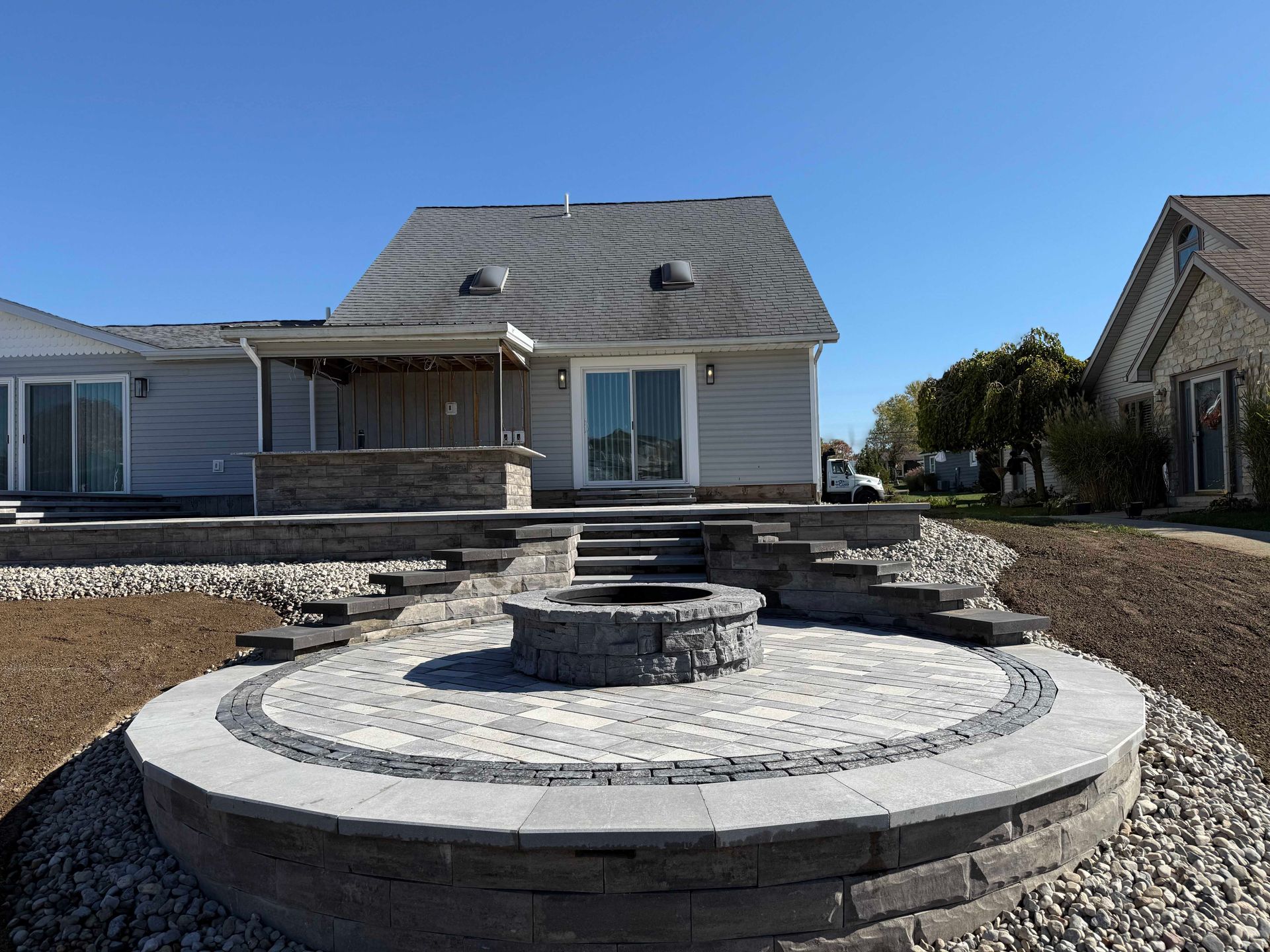 Backyard with fire pit, steps, and patio leading to a house with a gray roof and siding, on a sunny day.