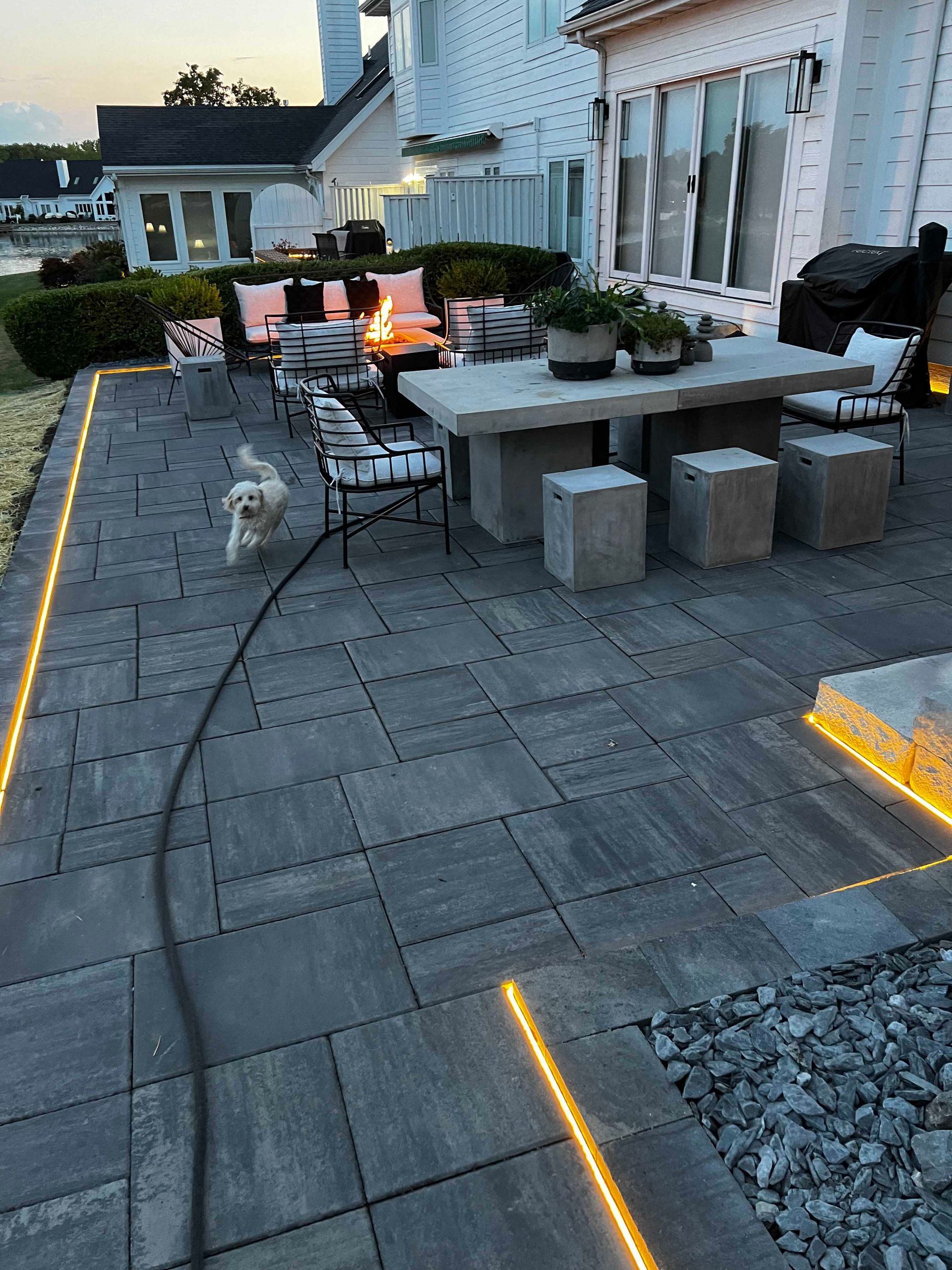 Patio with grey pavers, a stone table, seating, and a small dog on a leash.  Lit with warm, yellow lights at dusk.