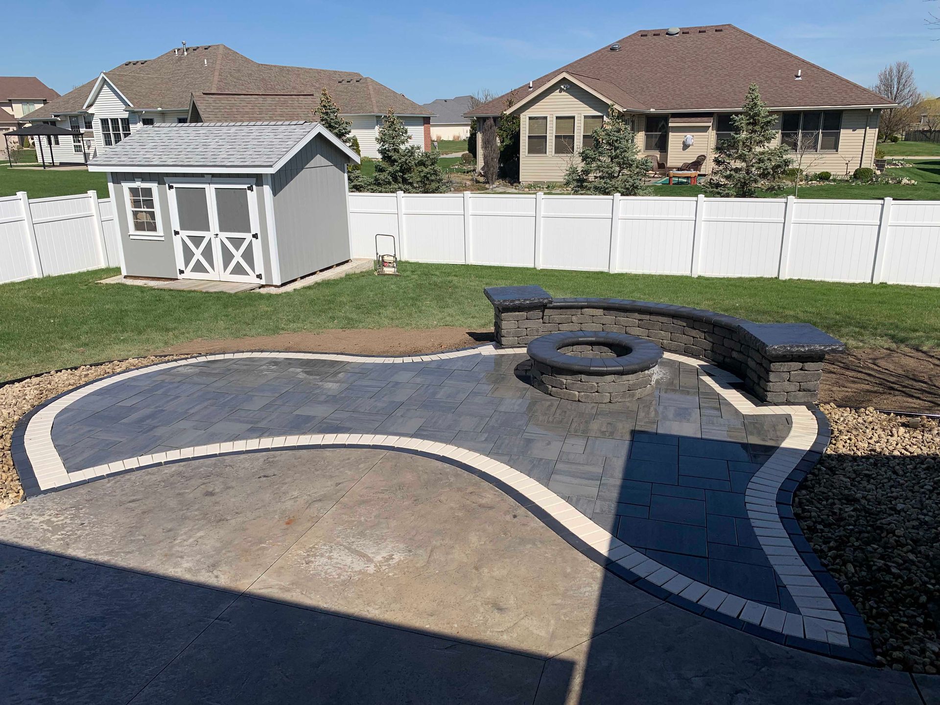 Backyard patio with fire pit and small gray shed, surrounded by white fence and grass.