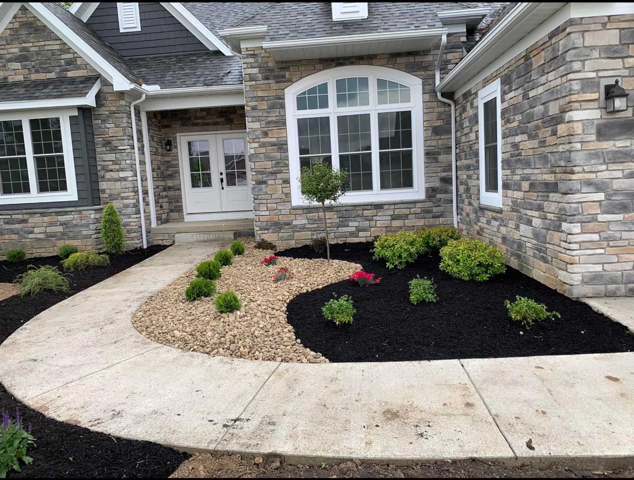 Stone house with a curved concrete walkway leading to the front door. Landscaping includes mulch and river rock beds.
