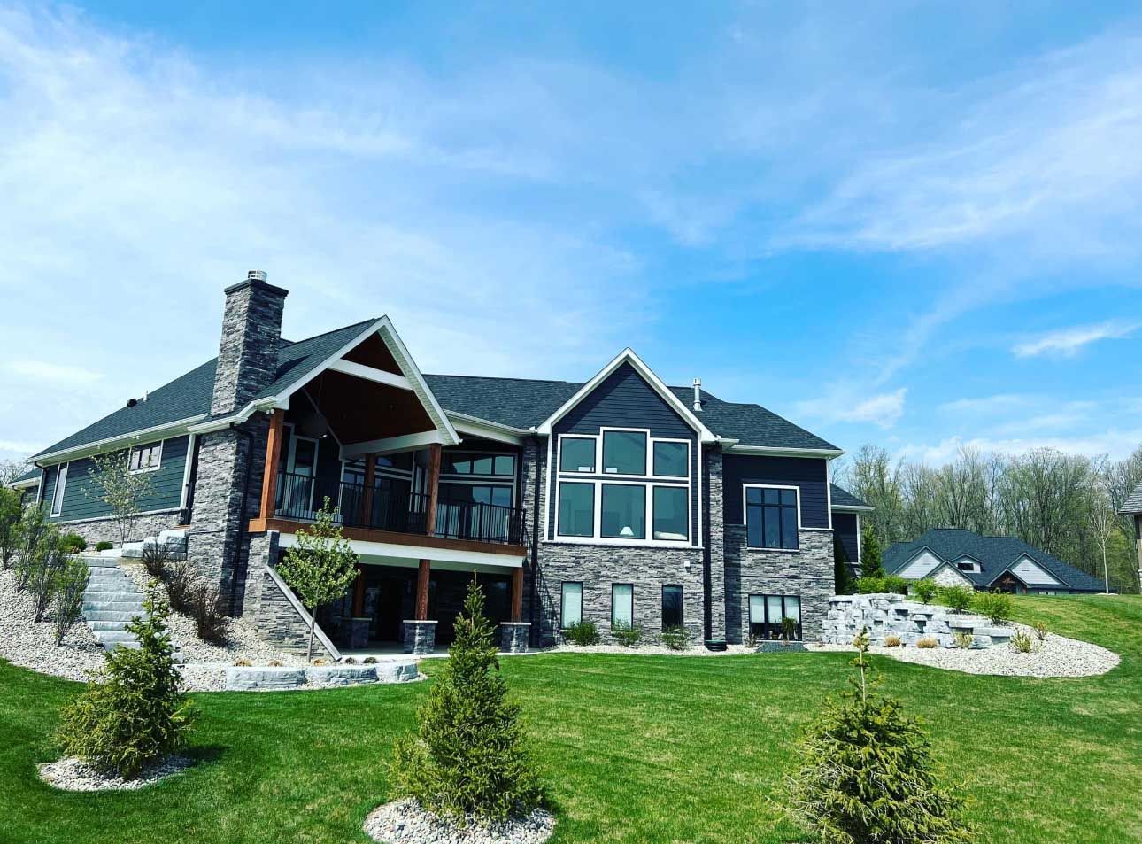 Large two-story house with dark gray siding, stone accents, and a covered deck under a bright blue sky.