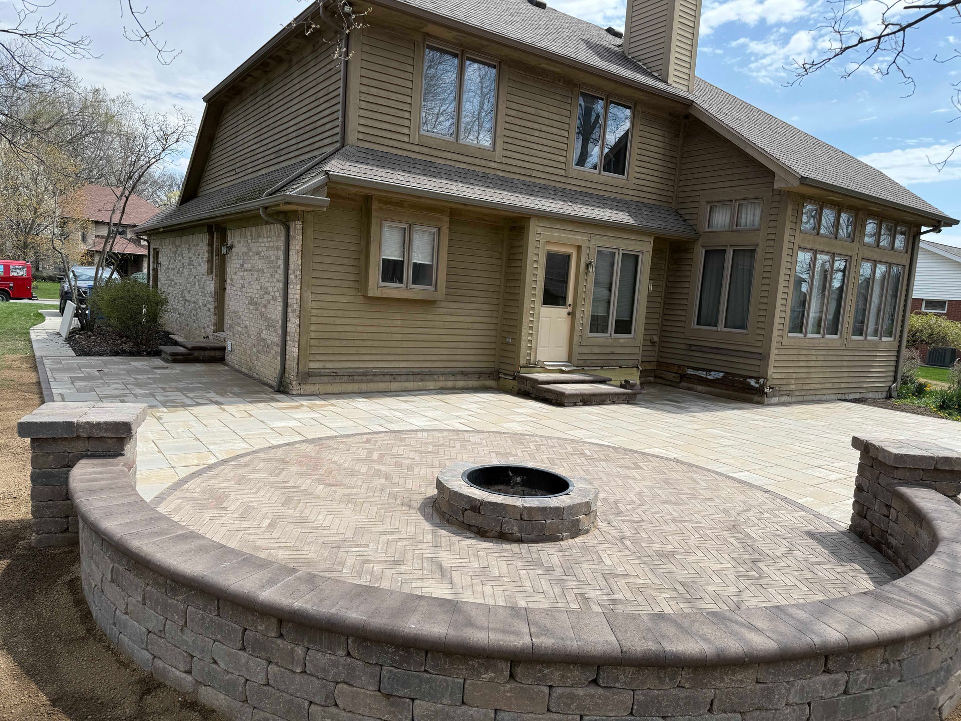 Backyard patio with fire pit, surrounding brick wall, two-story house with siding, clear skies.