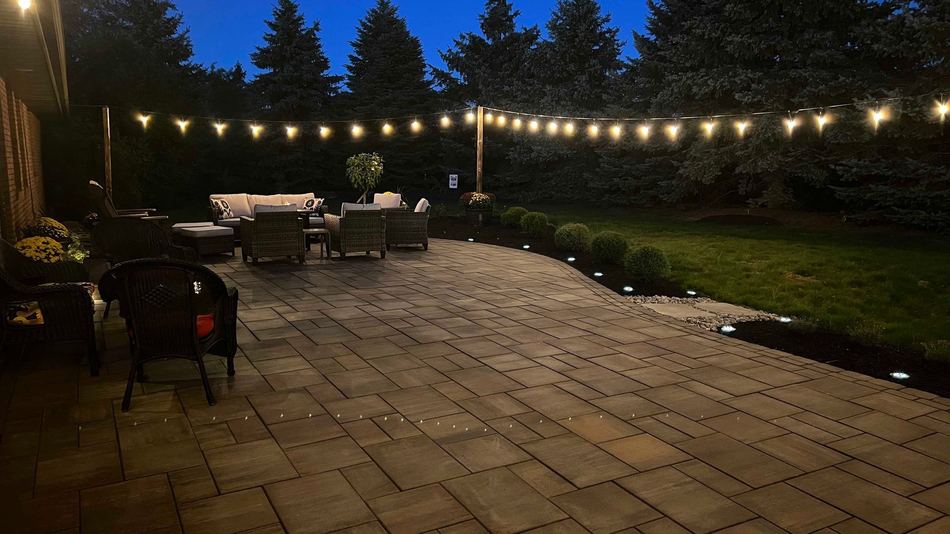 Patio lit with string lights at dusk, with outdoor furniture and a manicured lawn.