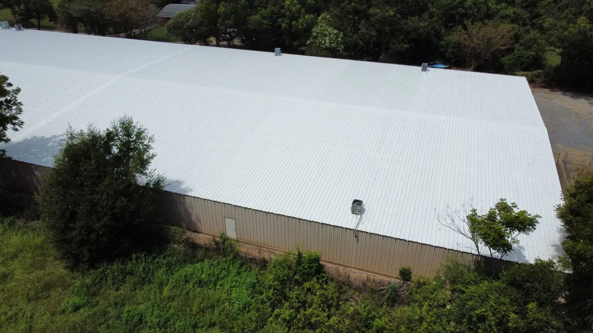 Aerial view of a building with a white metal roof, beige walls, and trees surrounding the structure.