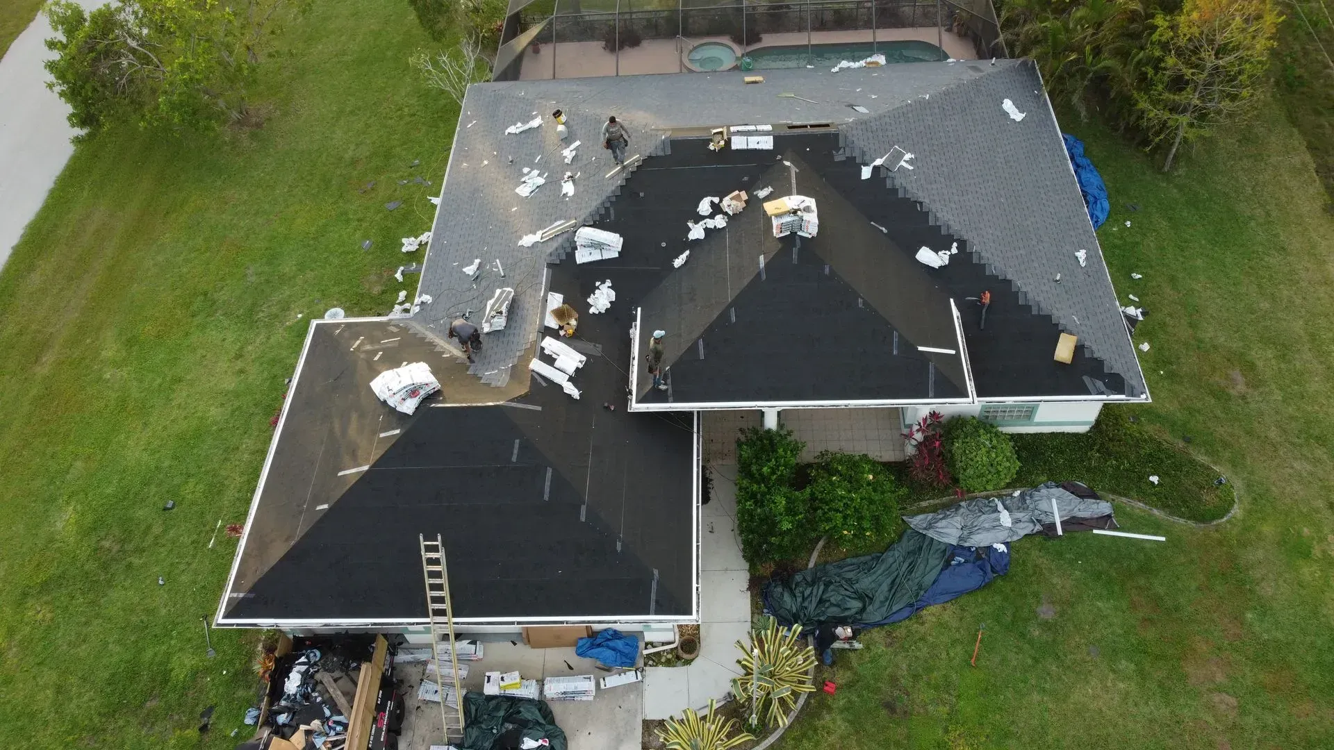 Roof of a house under construction with scattered materials on the black shingles. Green lawn surrounds the house.