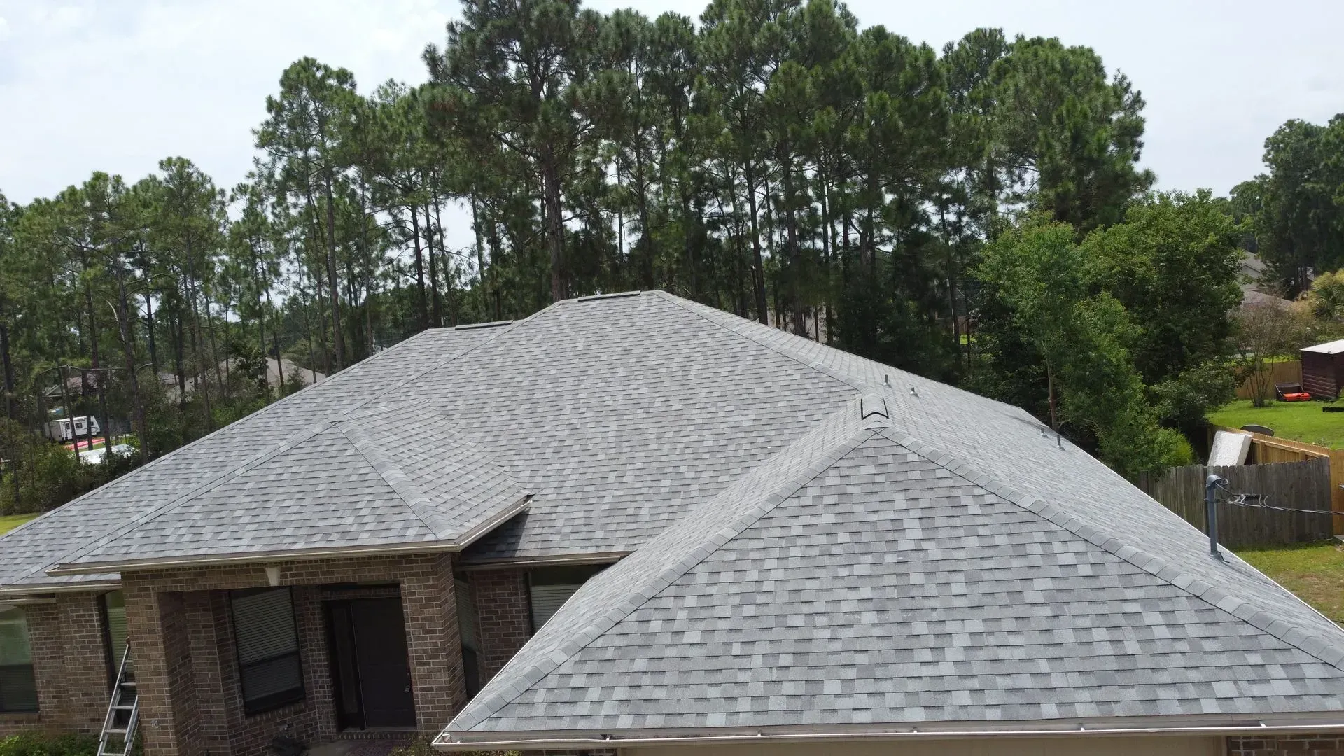 Gray shingle roof on a brick house with trees in the background under a cloudy sky.